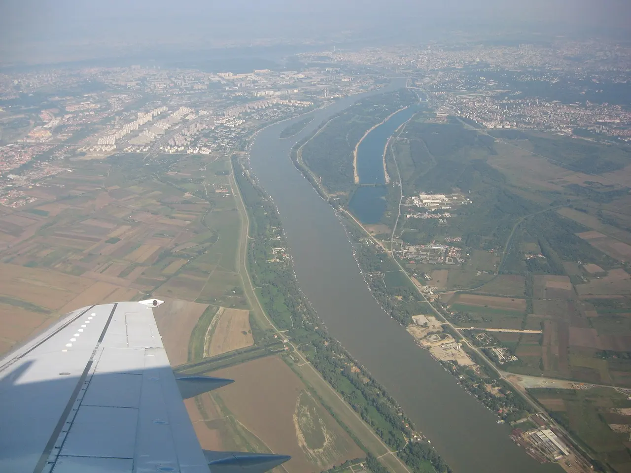 Aerial view of Ada Ciganlija as a wooded peninsula in the Sava, with the long dark rectangle of Lake Sava cut out of the river