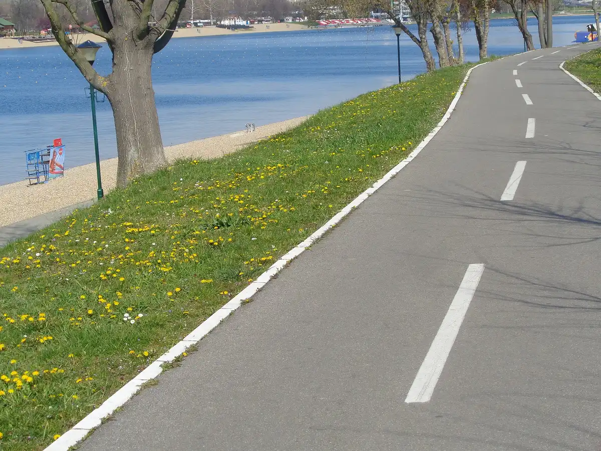 Paved two-lane path beside the pebble beach of Lake Sava, with a small lifeguard hut on the shore