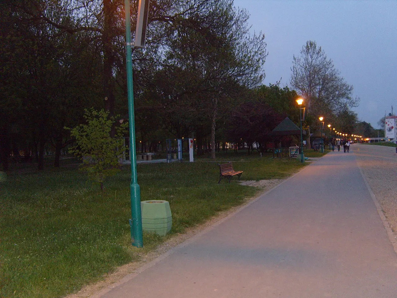 Lit asphalt promenade at Ada Ciganlija at dusk, with a park bench and outdoor exercise bars beside the path