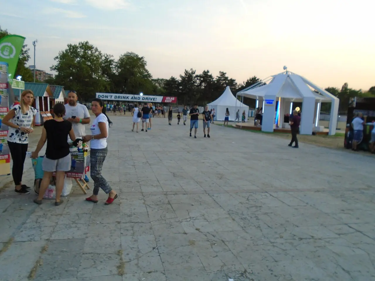 The paved plaza at Belgrade Beer Fest at dusk, festival-goers walking past a VIP tent and a Do not drink and drive sign