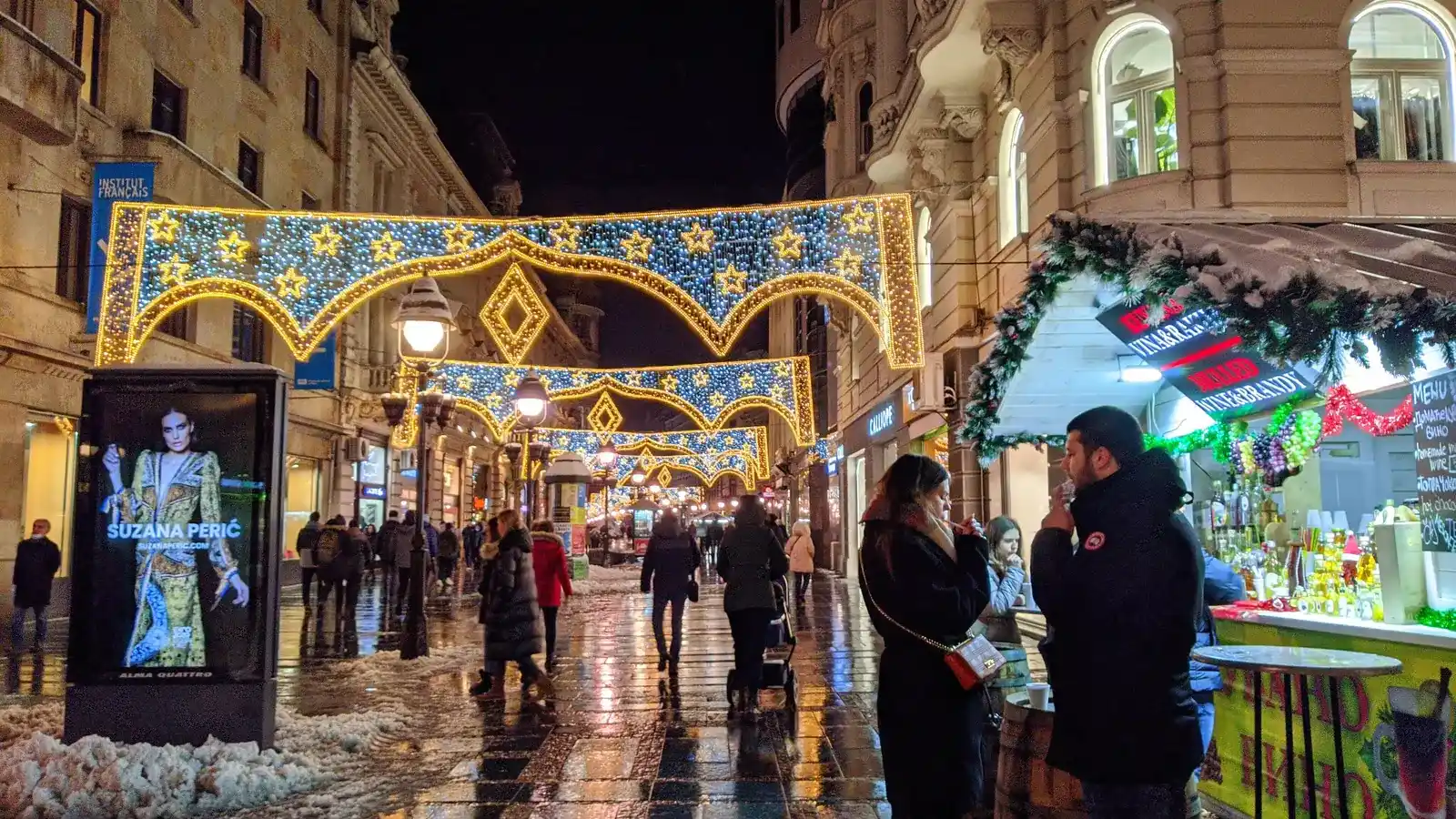 Knez Mihailova street in Belgrade at night under blue and gold light arches, with a mulled wine stall on the right and slush piled at the kerb