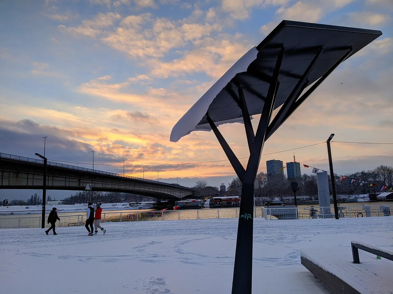 Snow lying on a Belgrade river quay at sunset, with a road bridge and the New Belgrade towers behind