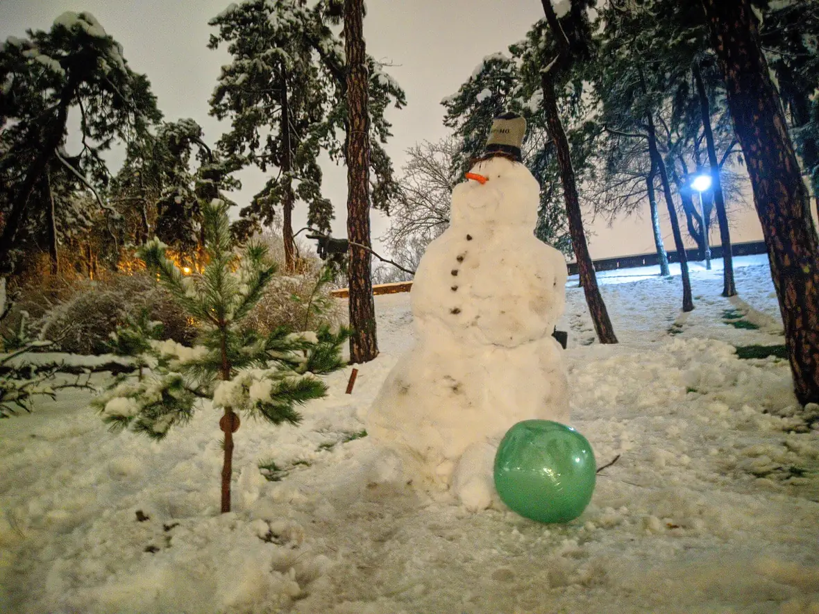 A lopsided snowman with a black hat standing among snow-covered pines in a Belgrade park at dusk