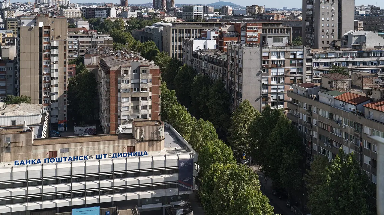 Belgrade rooftops with a Cyrillic bank sign and a green hill in the distance on a summer day