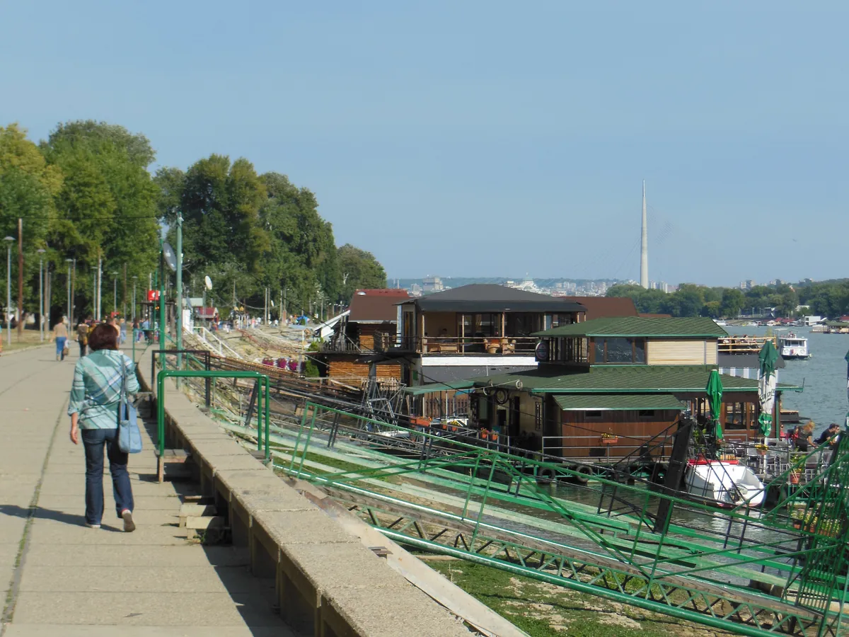 The Savski kej promenade in Belgrade with a line of moored floating bars along the bank and the Ada bridge pylon behind