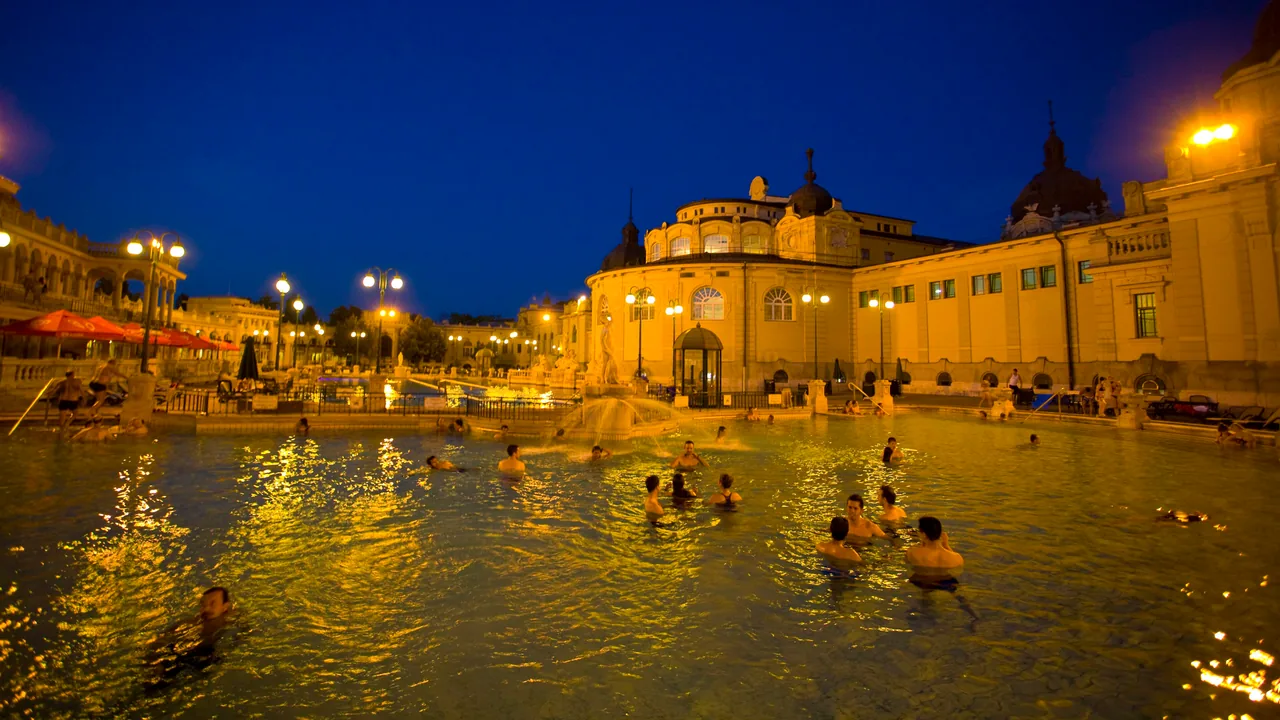 People in the floodlit outdoor pool of the Szechenyi thermal bath in Budapest at night, with the yellow bath building behind