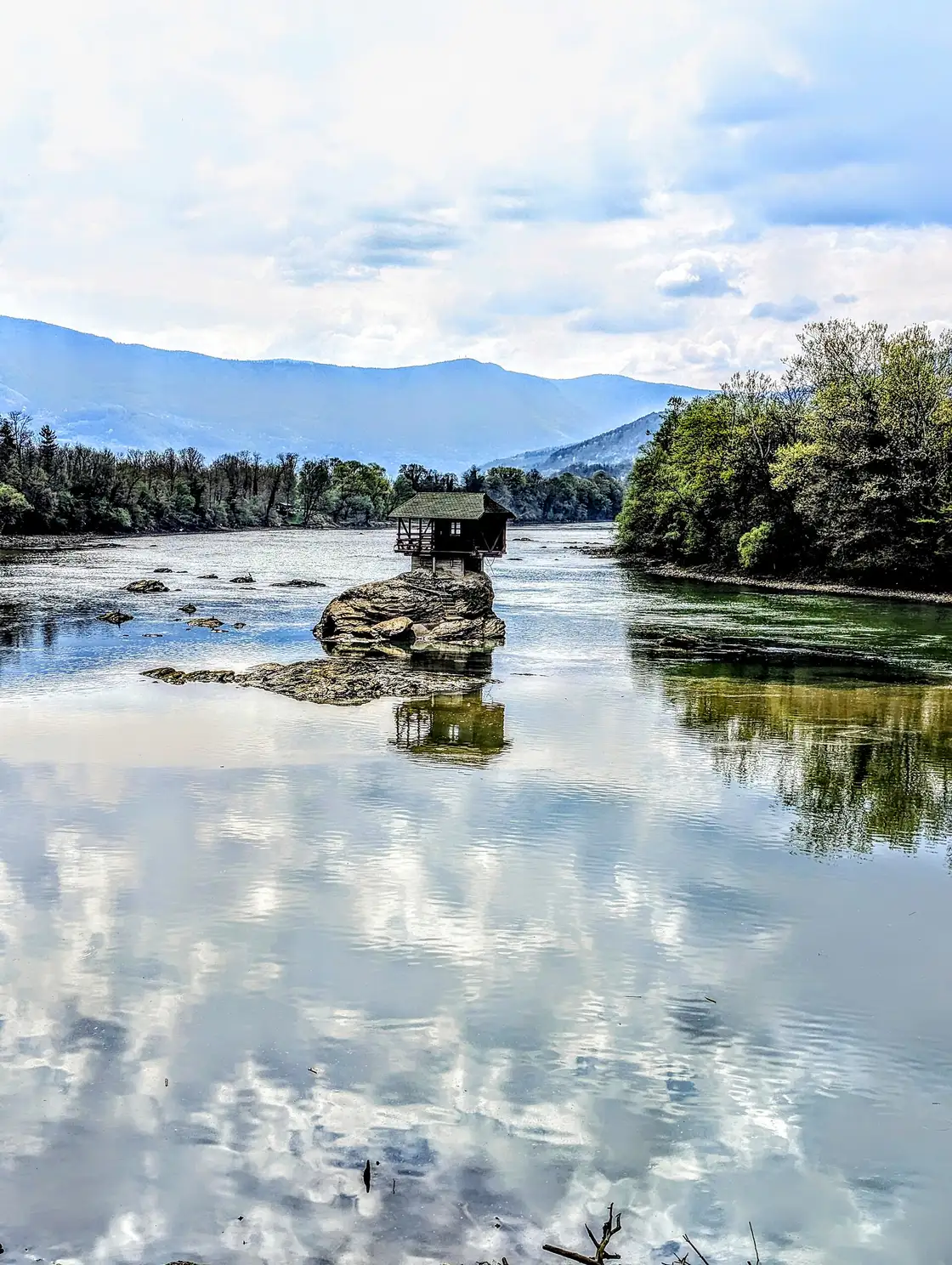 The Drina house in spring with the stone steps up the rock visible and mountains reflected in calm water