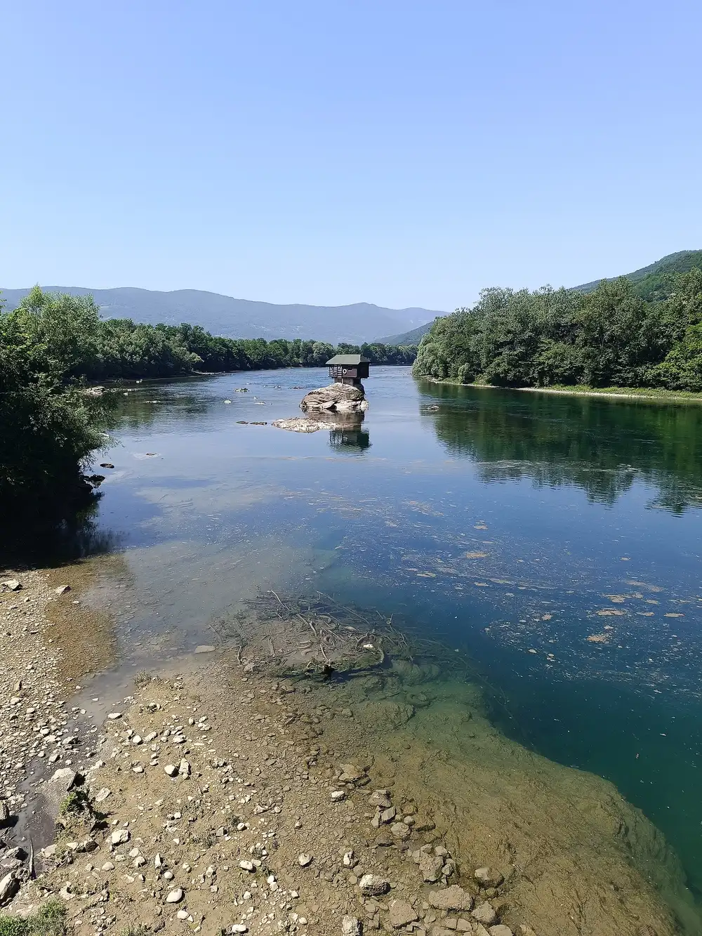 The Drina house on its rock under a clear blue sky with the river and pebble shore in the foreground
