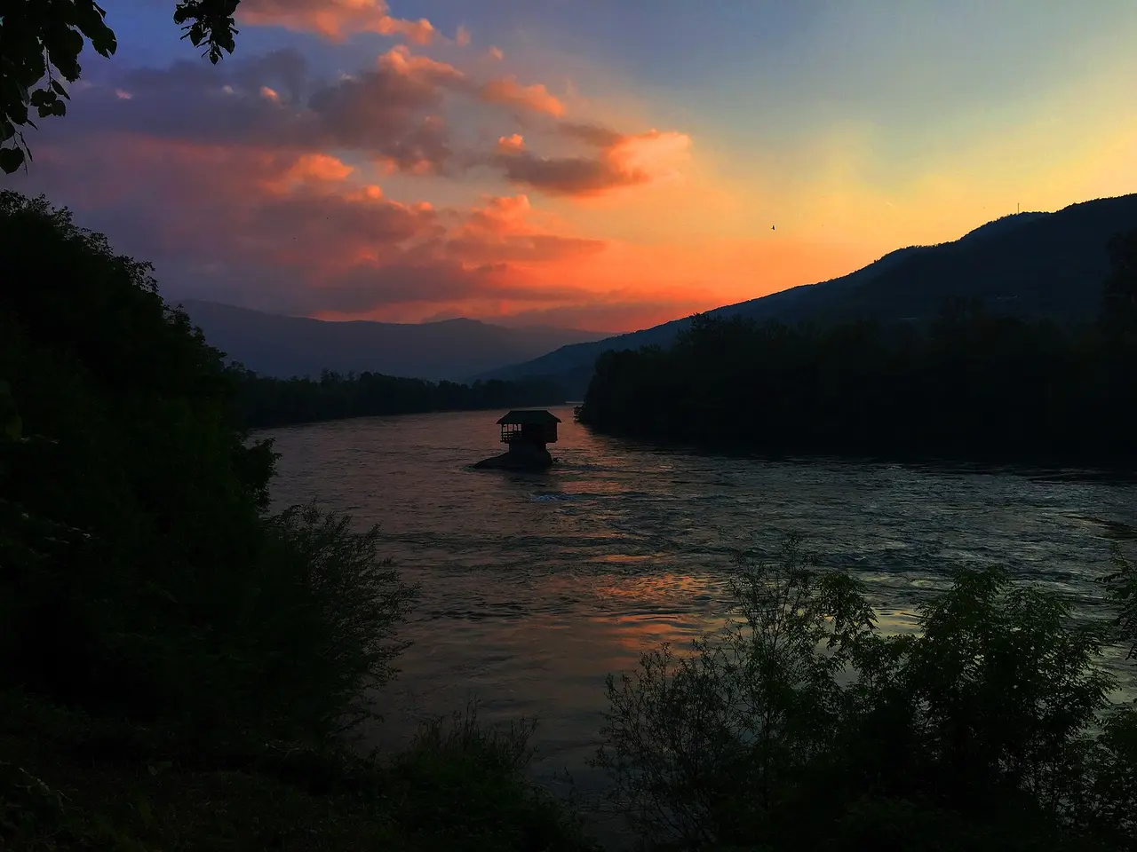 The Drina house silhouetted on its rock against an orange sunset over the river