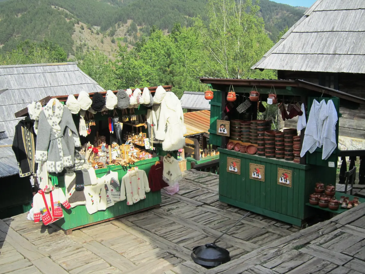 Two green wooden souvenir stalls in Drvengrad hung with knitted wool sweaters and stacked with clay pots