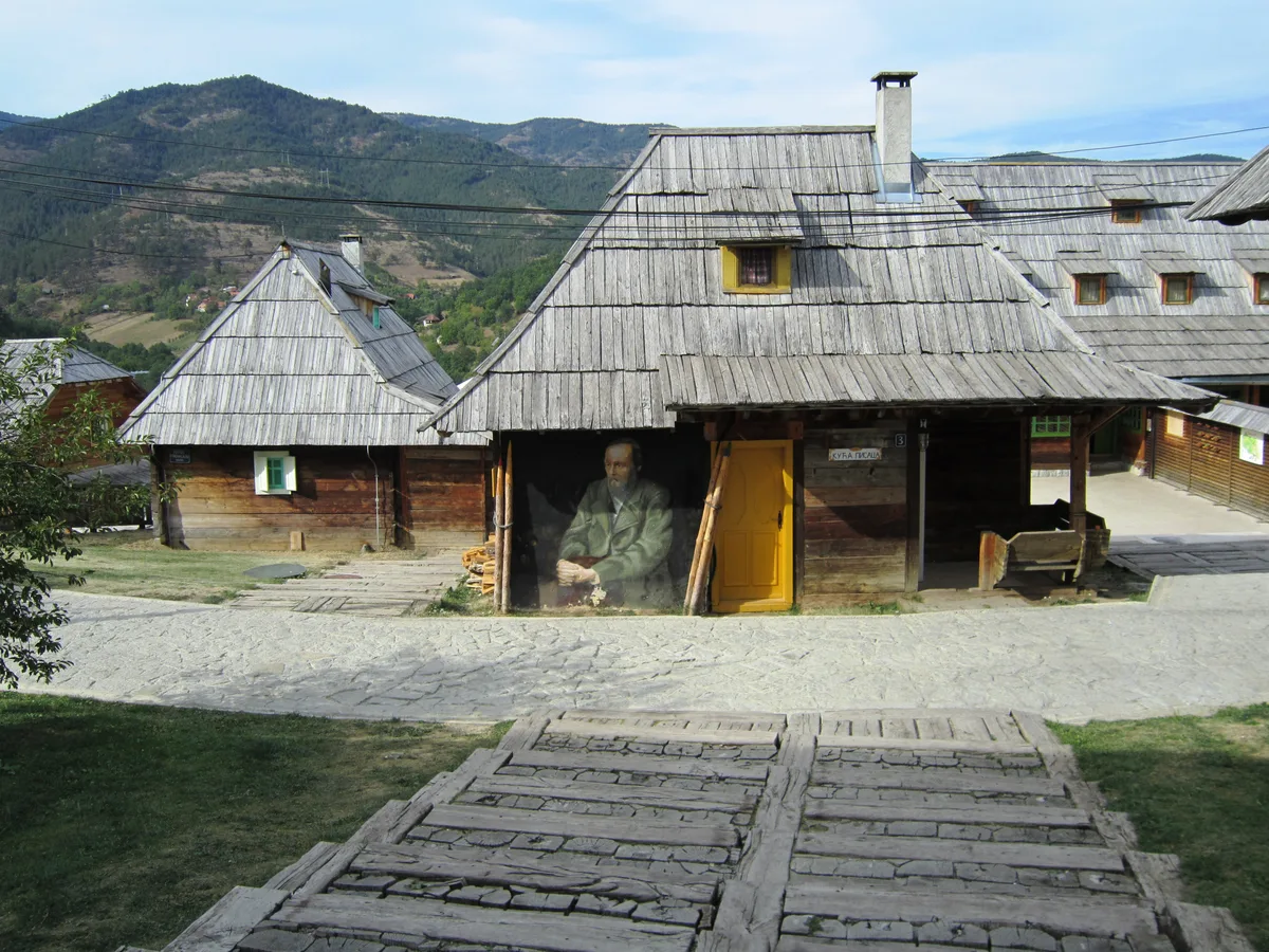 A large painted portrait mounted on the front of a timber house on the cobbled main square of Drvengrad, mountains behind