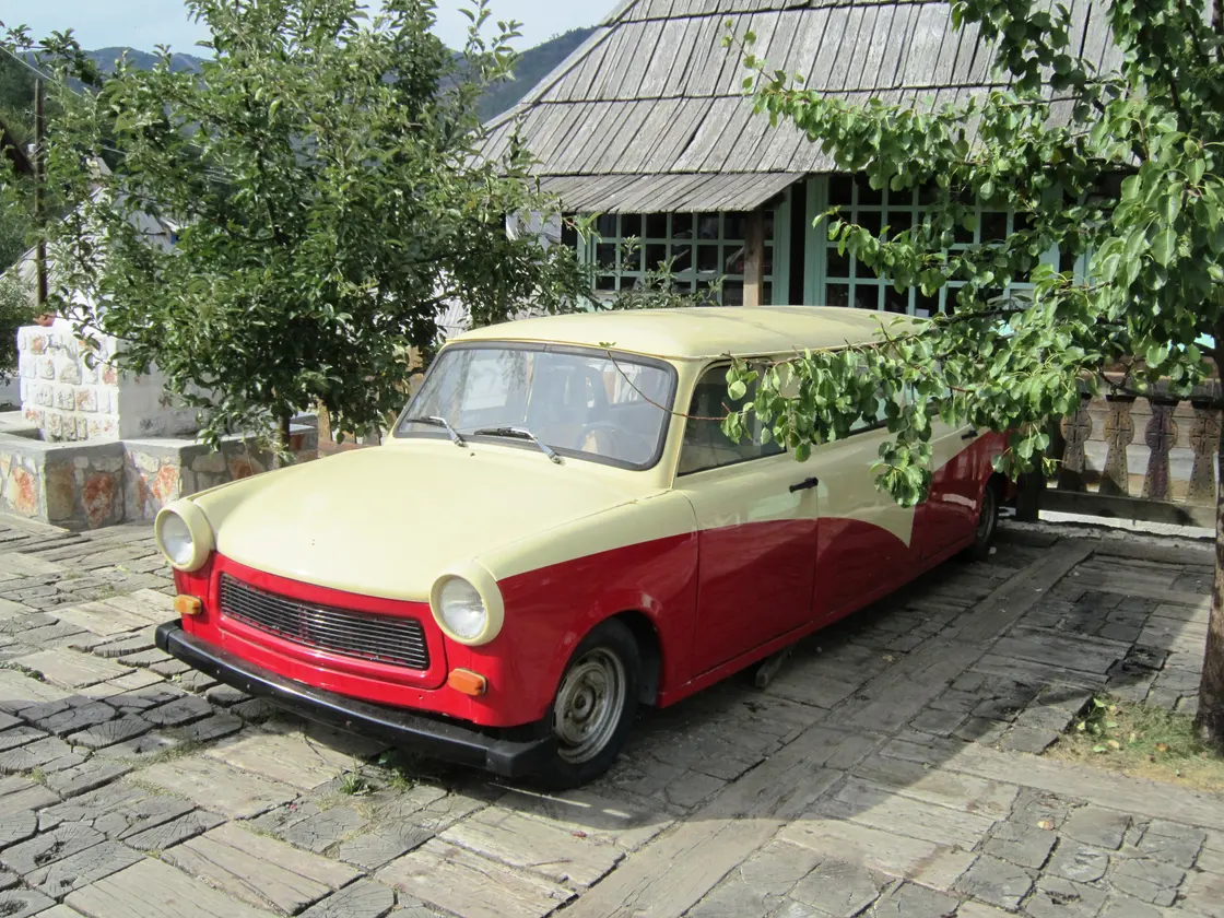 A stretched, elongated Trabant car in cream and red parked on the cobbles beside a timber house in Drvengrad