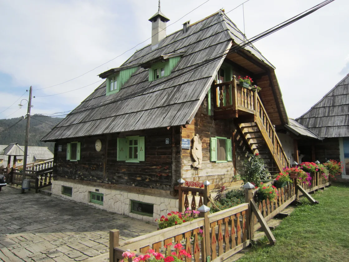 A traditional timber house in Drvengrad with green shutters, a shingled roof, an external wooden staircase and flowers