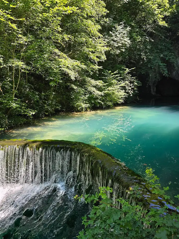 The emerald-turquoise water of the Krupaj spring below a small weir and forested cliff