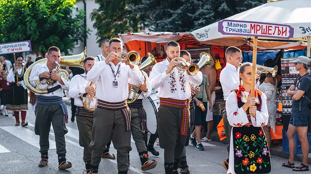 A brass band in white folk shirts marching and playing trumpets through Guča, a girl in embroidered costume carrying the band sign