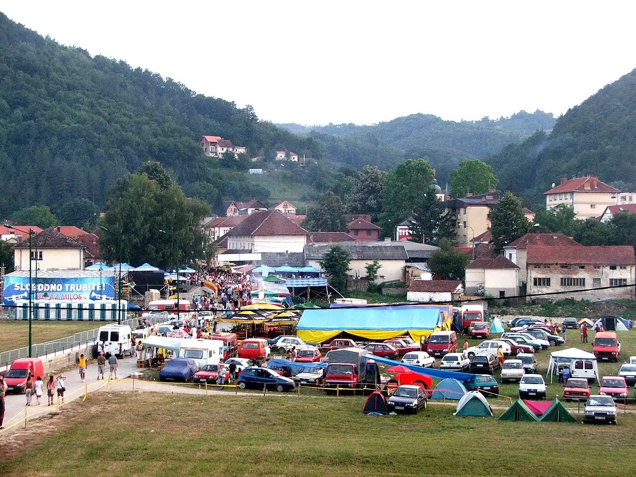 The Guča festival grounds seen from above: tents, parked cars in the fields and market stalls with the village and green hills behind