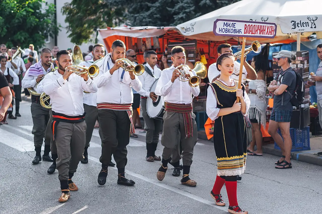 A competing trumpet orchestra playing in the street at Guča, led by a young woman in traditional dress holding the orchestra name board