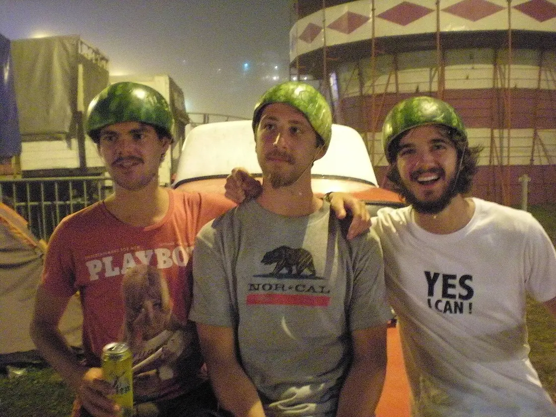 Three young festival-goers at night wearing hollowed-out watermelon rinds as helmets, one holding a beer, grinning