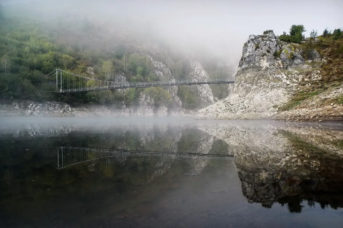 A footbridge and misty limestone cliffs reflected in the still water of the Uvac river in western Serbia