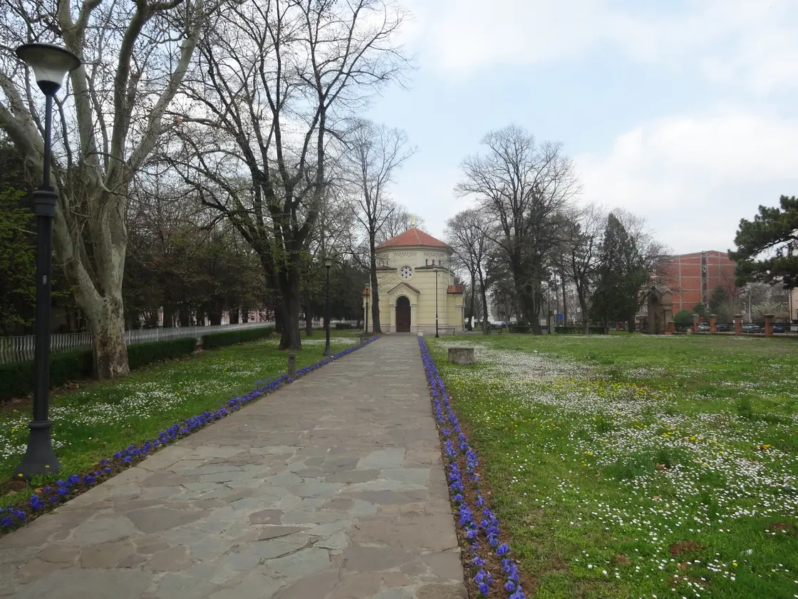 The small yellow chapel that encloses the Skull Tower in Niš, seen down a park path lined with spring flowers