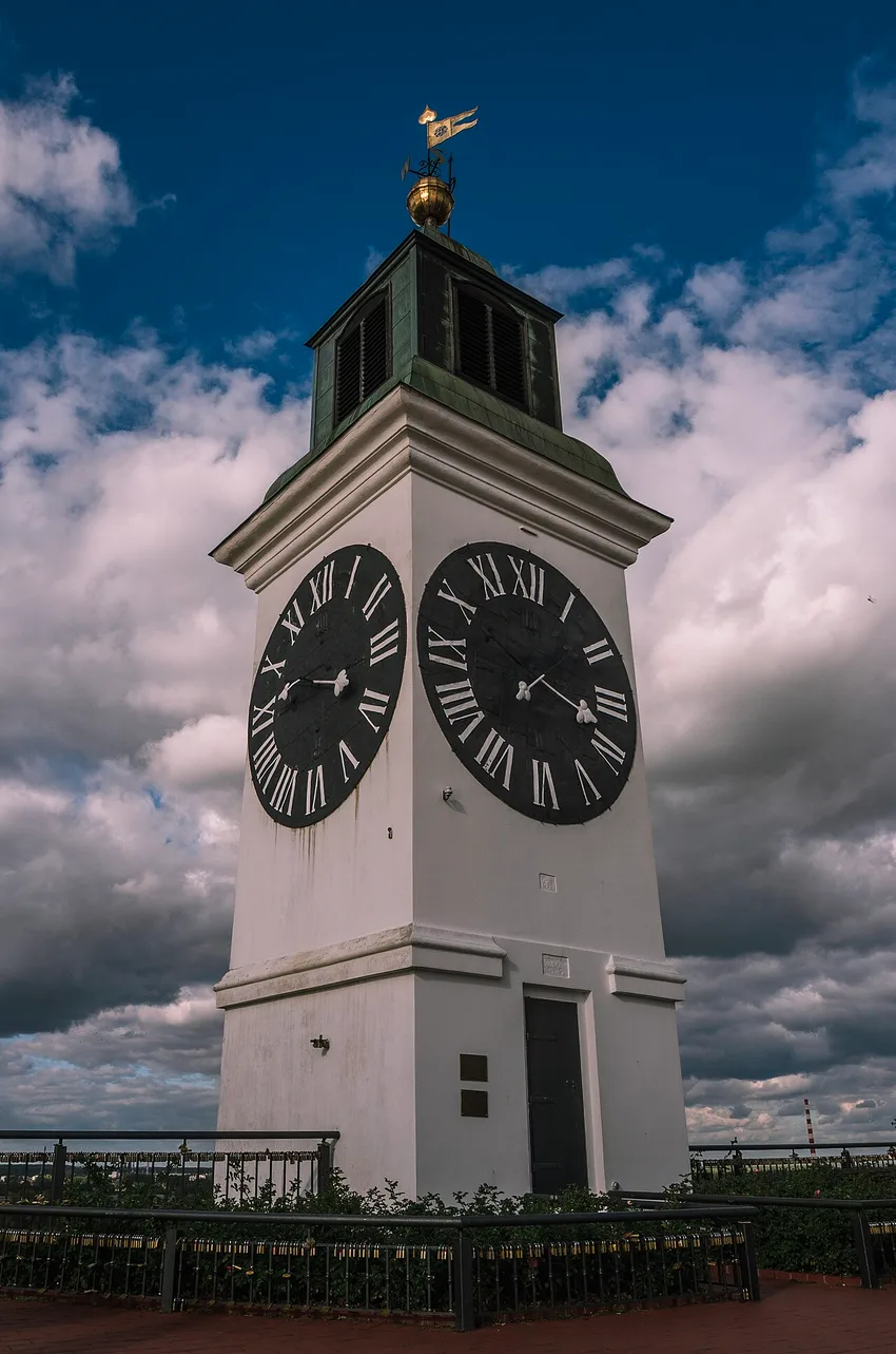 The white Petrovaradin clock tower against a dramatic sky, its two black faces showing the oversized hour hand and small minute hand