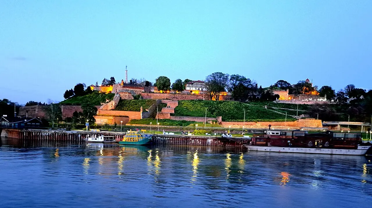 Belgrade Fortress and the lit Pobednik monument on the hill above the river at dusk, barges moored below