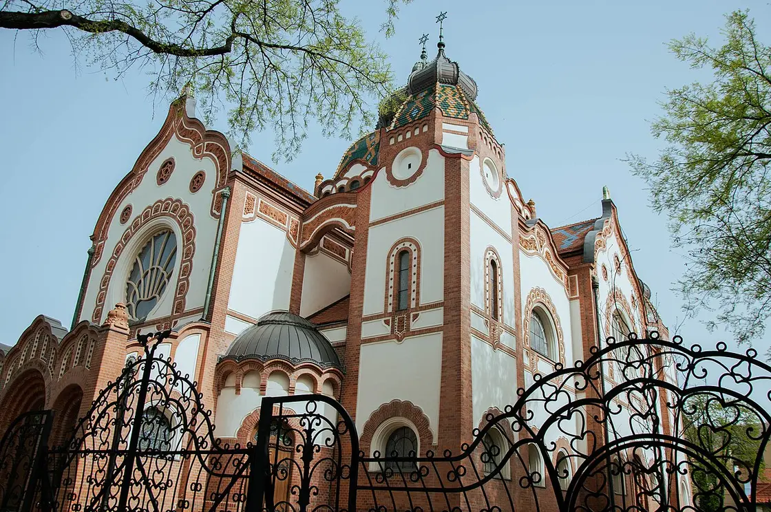 The white and red-brick Art Nouveau synagogue of Subotica with its green and gold tiled dome under a clear sky