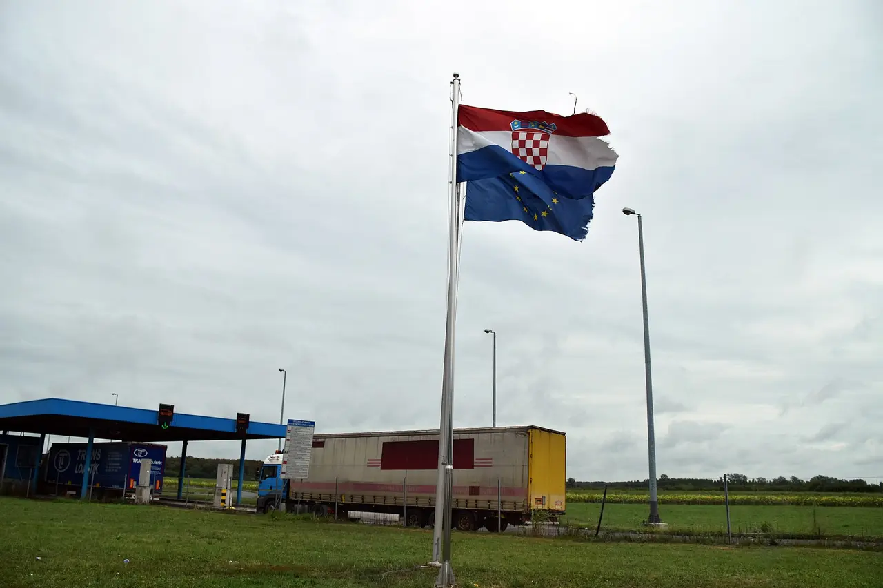 A frayed Croatian flag and EU flag flying over the Bajakovo crossing with a lorry at the canopy behind