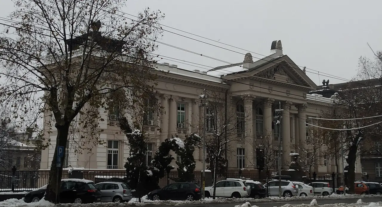 The columned facade of the Svetozar Markovic University Library in Belgrade behind bare trees and parked cars in the snow