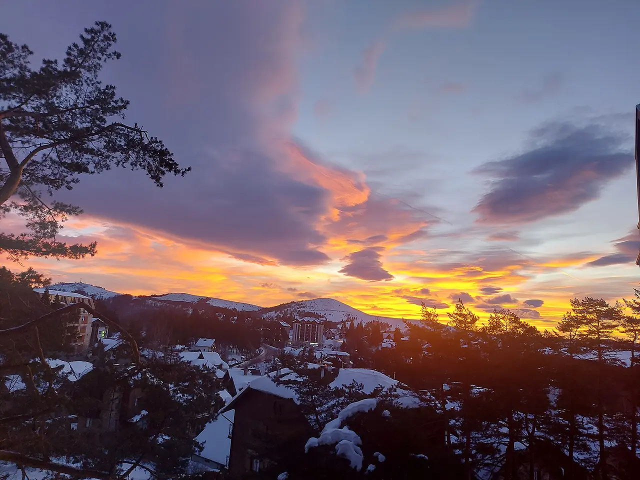 Snow-covered rooftops and pine trees on Zlatibor under an orange and purple sunset sky