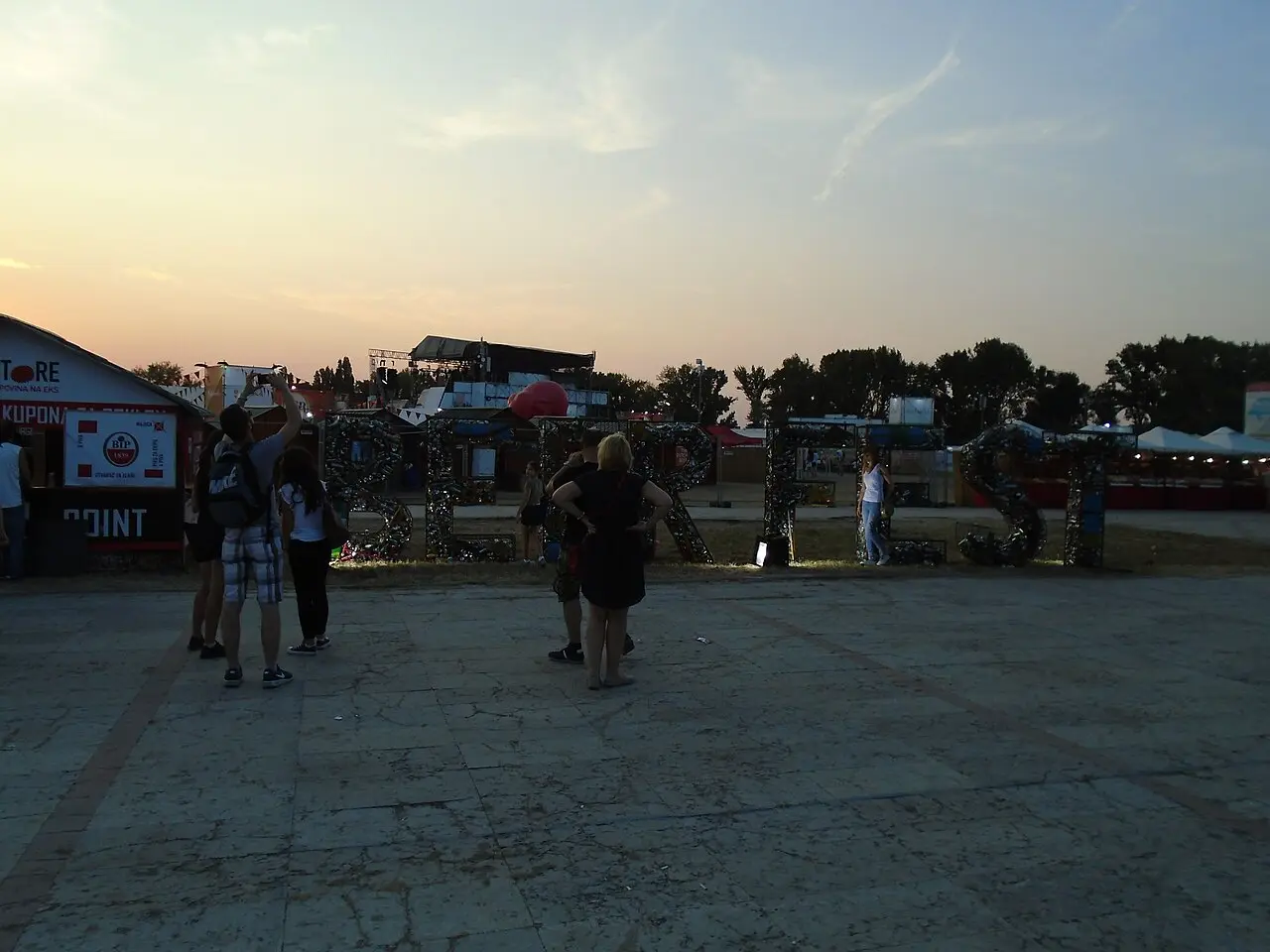 The grounds of Belgrade Beer Fest at dusk, crowds by the big BEER FEST letters made of bottle caps with a stage behind