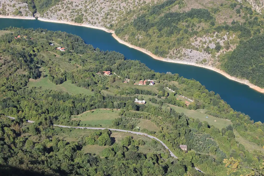 The winding turquoise Zavoj reservoir seen from above, with scattered farmhouses on the green slopes around it