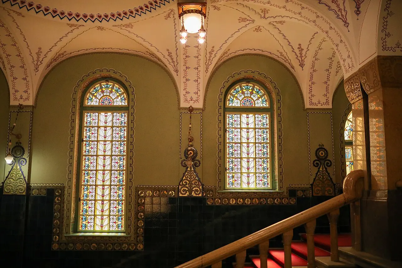 A stairwell inside Subotica City Hall with tall stained-glass windows and painted vaults