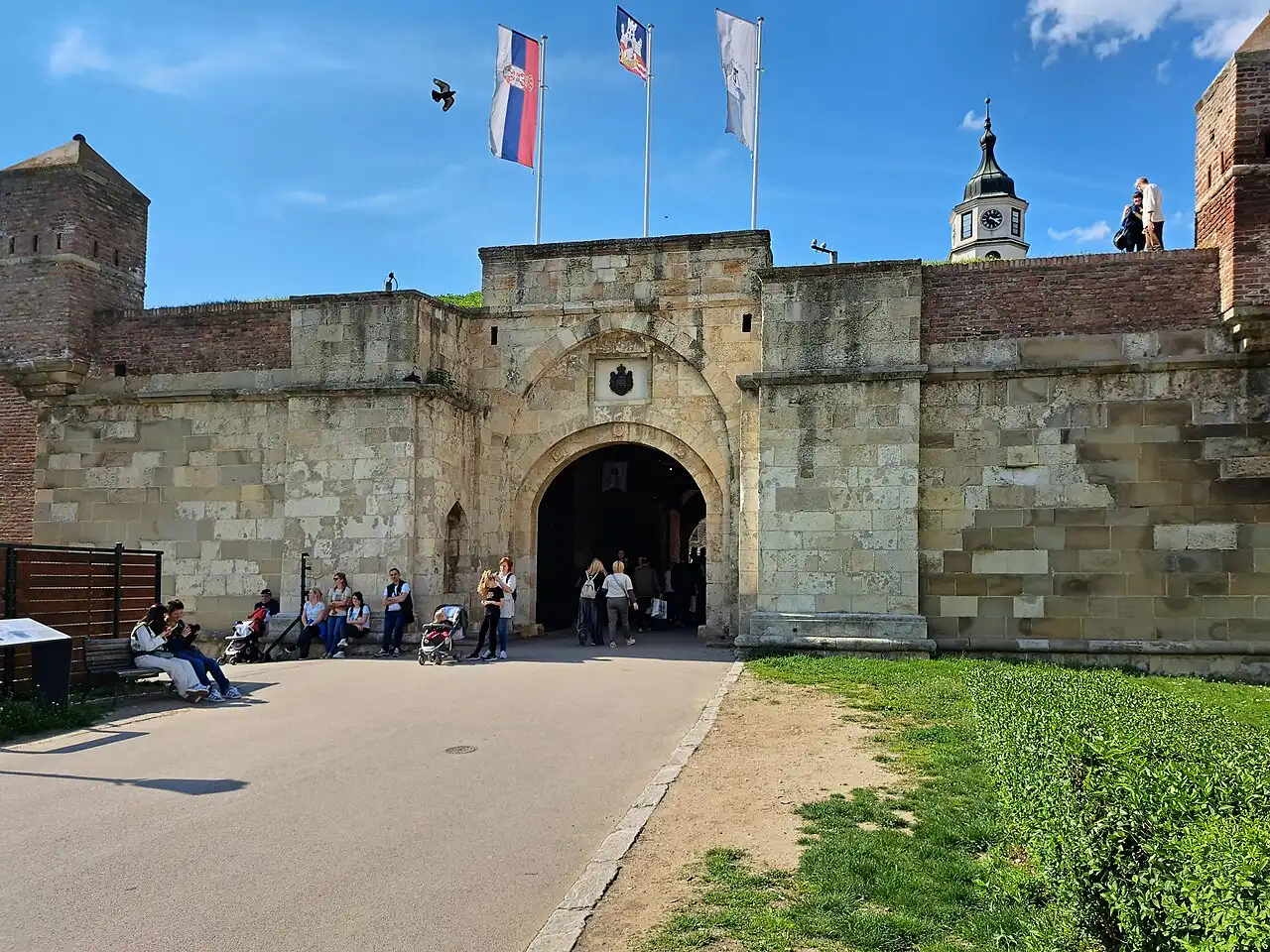 A stone gate in the ramparts of Belgrade Fortress with Serbian flags flying above and the clock tower behind
