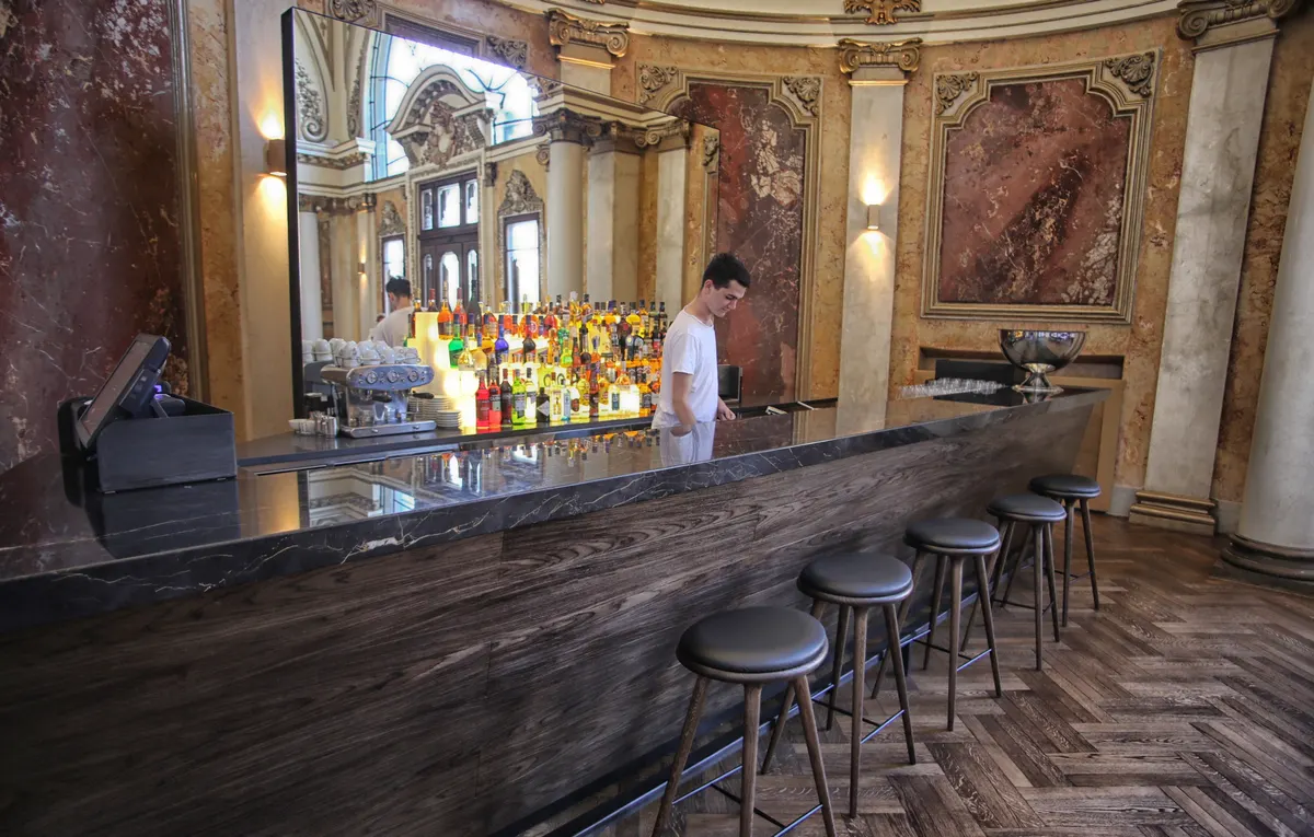 A bartender behind a marble counter in an upscale Belgrade bar, with a card payment terminal on the counter