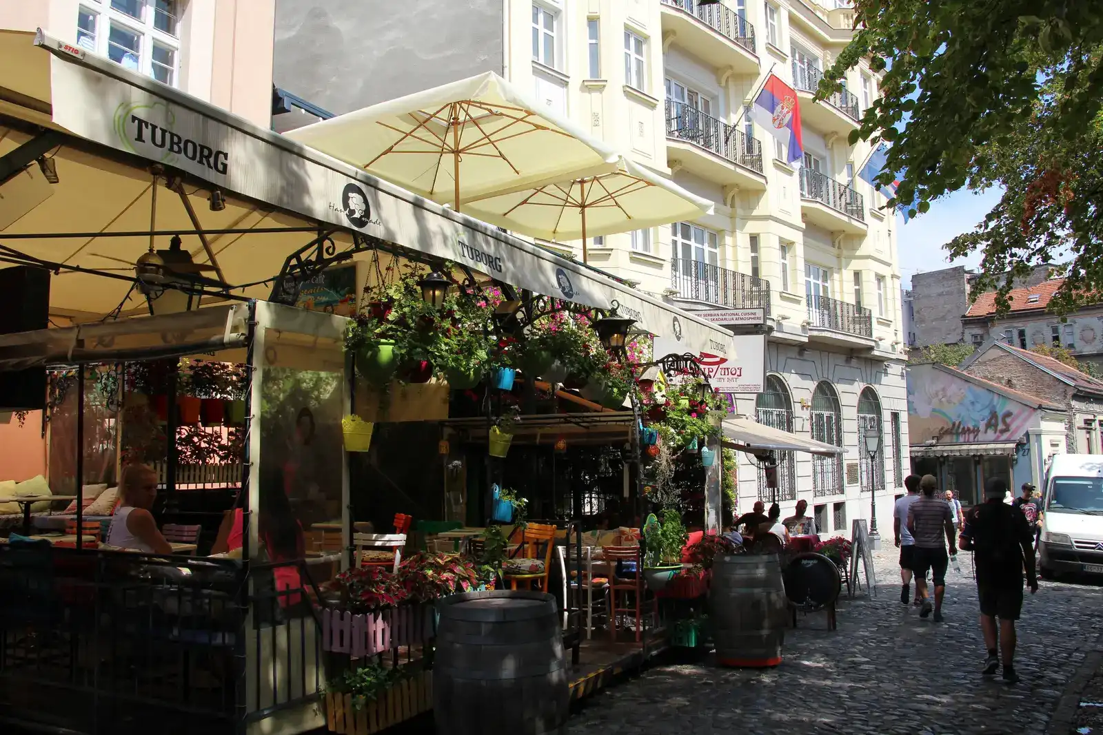 A kafana terrace in Skadarlija, Belgrade's old bohemian street, with outdoor tables under umbrellas and a Serbian flag on the building behind