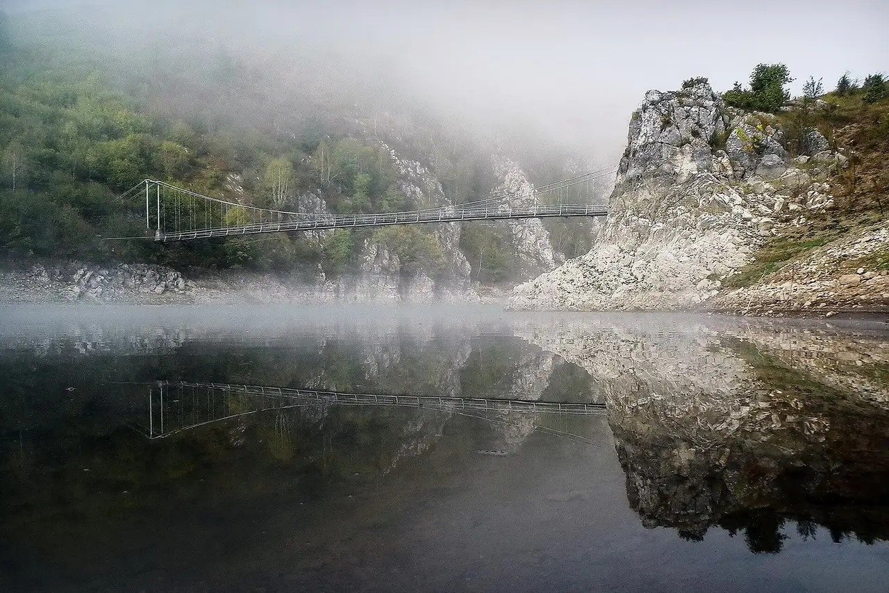 A suspension footbridge over the still water of the Uvac reservoir in morning mist, reflected below