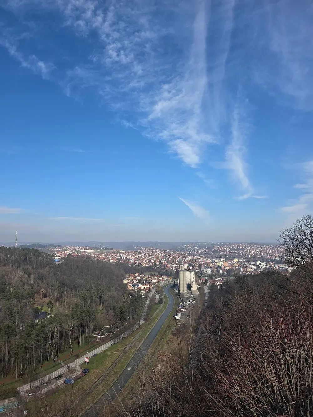 A winter view over Valjevo from a hill above the town, showing the valley, a channelled stream and forested slopes beyond