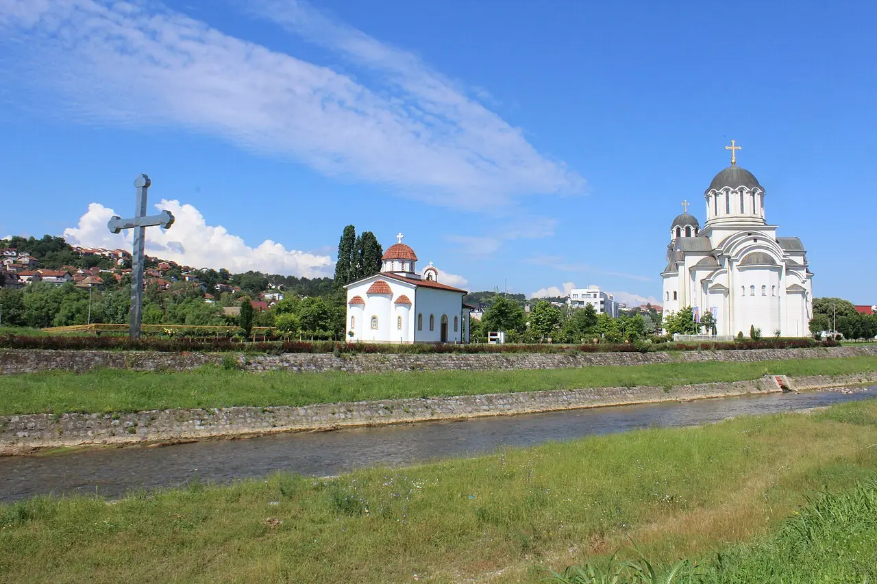 The Church of the Resurrection of Christ in Valjevo, a large white cathedral seen across a meadow with a memorial cross and a smaller church in front of it