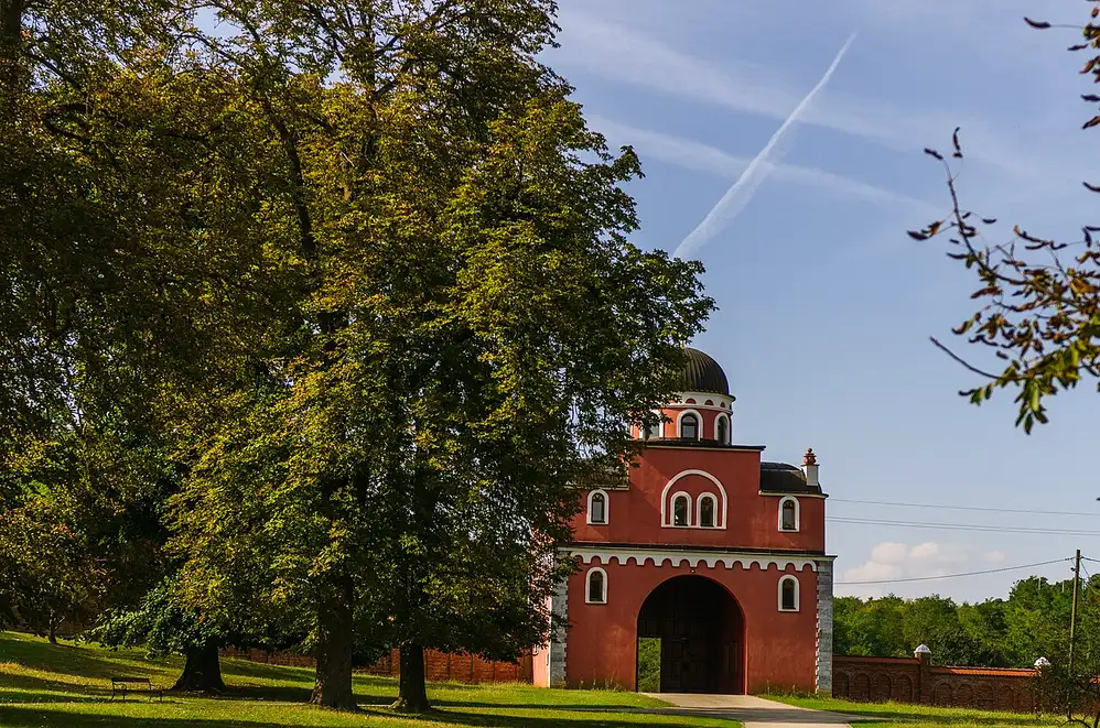 The red gate tower and dome of Krušedol monastery framed by tall trees on Fruška Gora