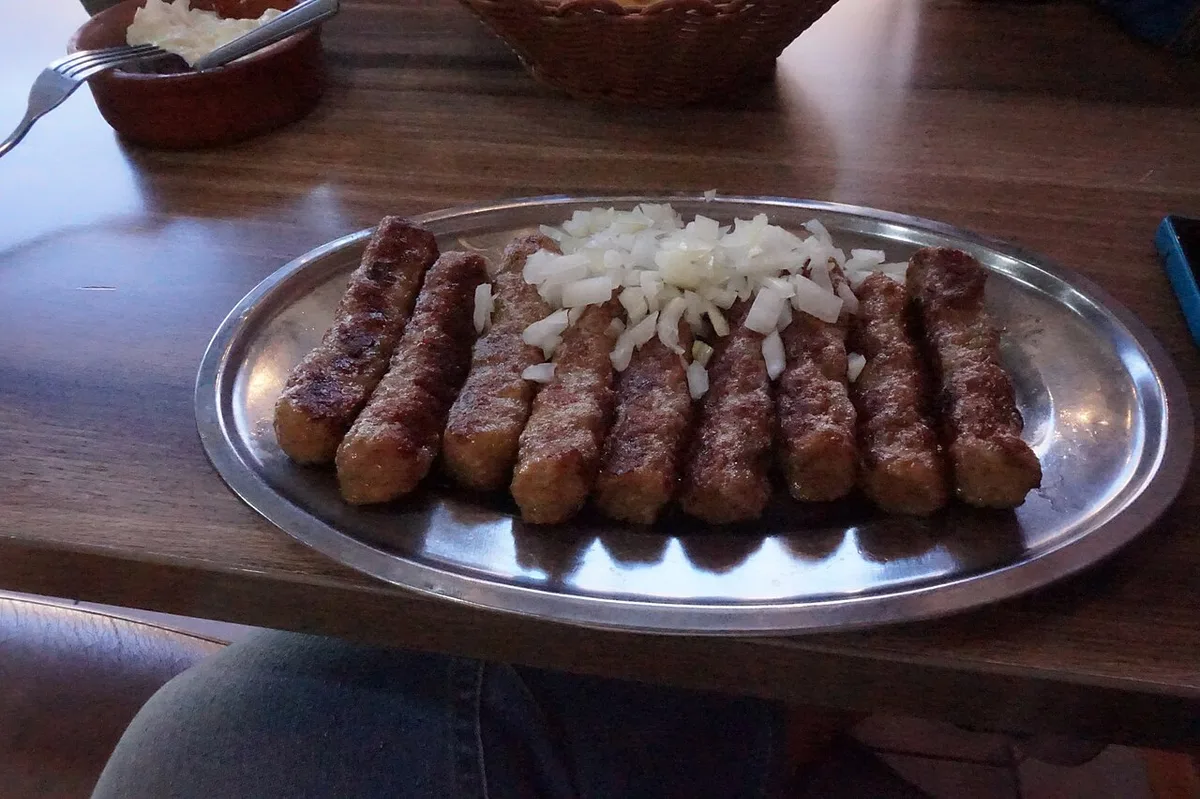 A metal platter of grilled ćevapi meat rolls topped with chopped raw onion, served in a Serbian grill house