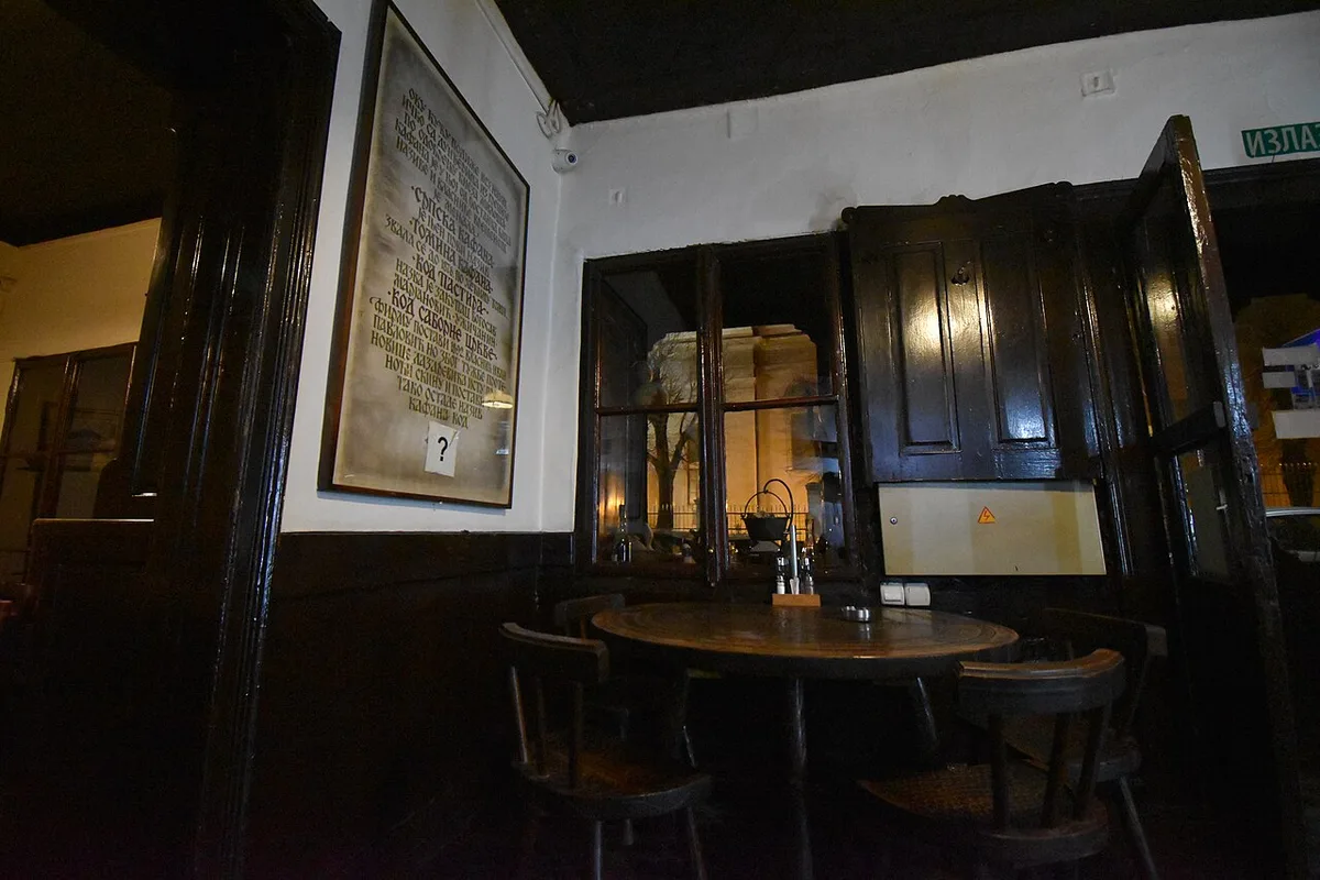 The dark wood interior of Znak Pitanja, an old Belgrade kafana, with round tables and an old framed text on the wall
