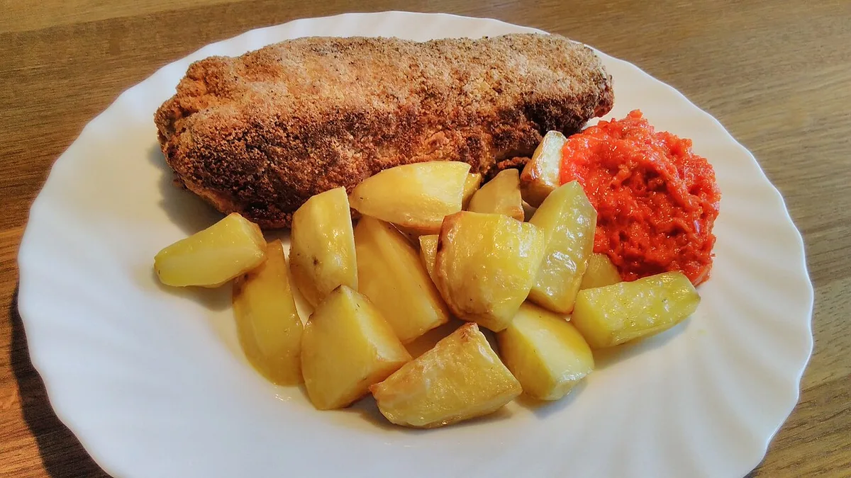 Karađorđeva šnicla, a golden breaded rolled cutlet, on a white plate with roast potatoes and a spoon of red ajvar