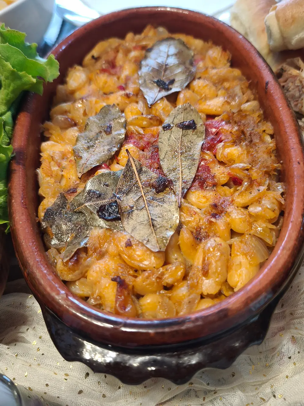 A clay pot of baked prebranac beans topped with bay leaves, on a laid Serbian celebration table
