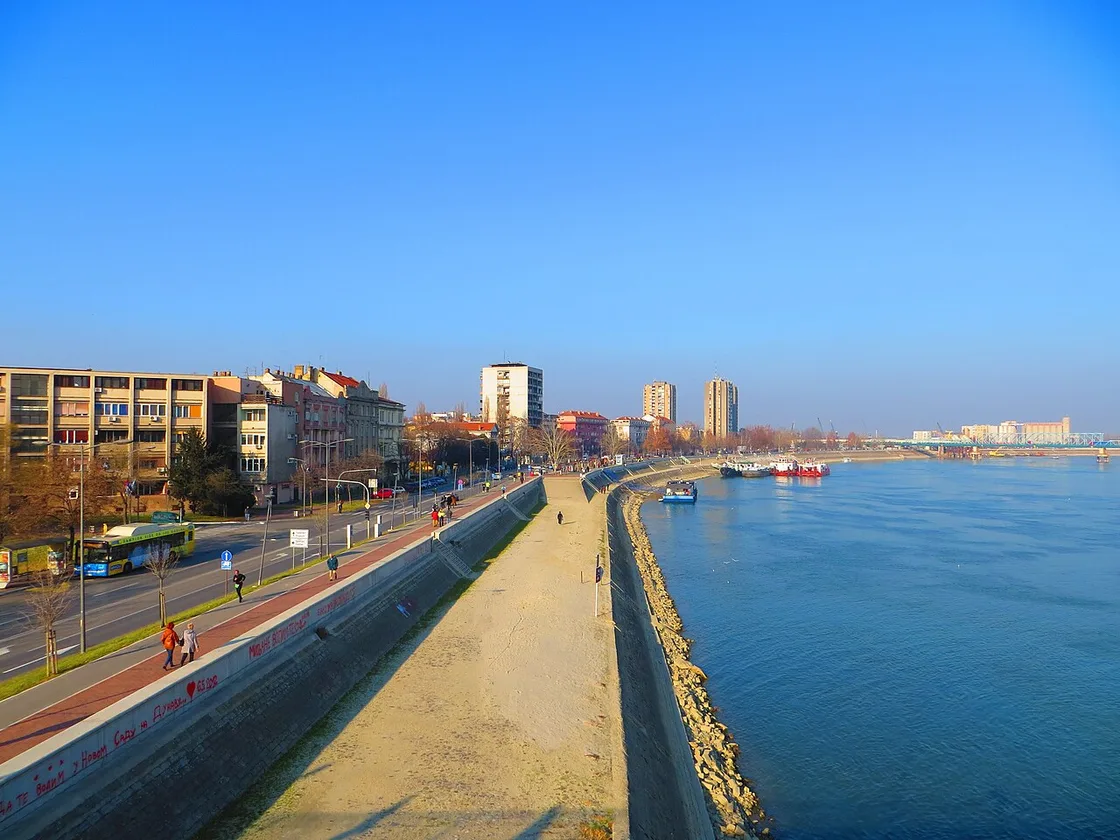 The Danube embankment in Novi Sad with a riverside road, apartment blocks and moored boats on a clear day